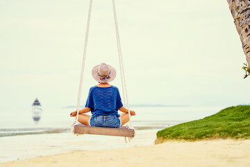 Vacation concept. Happy young woman in hat sitting on swing enjoying sea view.