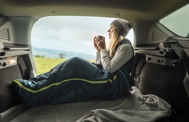 Girl resting in her car. Woman hiker, hiking backpacker traveler camper in sleeping bag, relaxing, drinking hot tea on top of mountain. Road trip. Health care, authenticity, sense of balance.