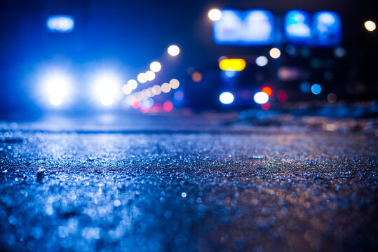Rainy night in the big city, glare from the headlights of the parked car and a passing near the stream of cars. Close up view from the sidewalk level