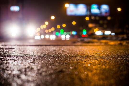 Rainy night in the big city, glare from the headlights of the parked car and a passing near the stream of cars. Close up view from the sidewalk level