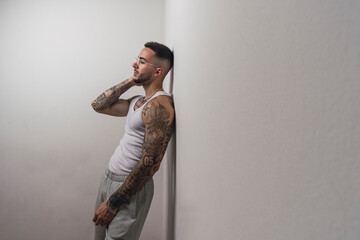 Retrato de un joven apuesto varón español tatuado posando apoyado en una pared blanca