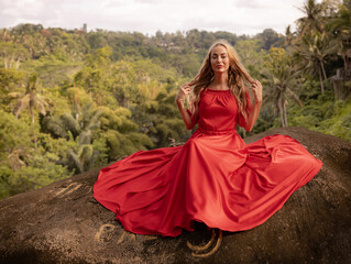 Obraz na płótnie Canvas Bali trend photo. Caucasian woman in long red dress sitting on big stone in tropical rainforest. Vacation in Asia. Travel lifestyle. Breathtaking view. Bongkasa, Bali, Indonesia