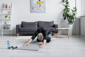 man with grey hair stretching on fitness mat near dumbbells in living room, banner.