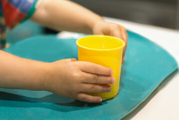 Hands of a child with a mug.A cute little girl holds a plastic yellow mug of warm milk in her hands for a morning breakfast at home, sitting at the dining table, close-up. Soft Focus.Healthy lifestyle