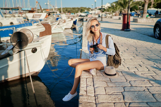 Photography and travel. Young woman with rucksack holding camera sitting on wharf with beautiful sea view with fishing boats.