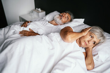 Obraz na płótnie Canvas Senior couple in the bed. Pretty aged woman is angy with her sleeping husband covering ears with hands.