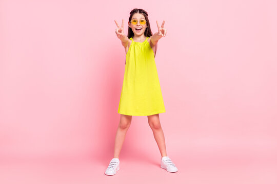 Full length photo of funny adorable schoolgirl dressed yellow outfit dark glasses showing two v-signs smiling isolated pink color background