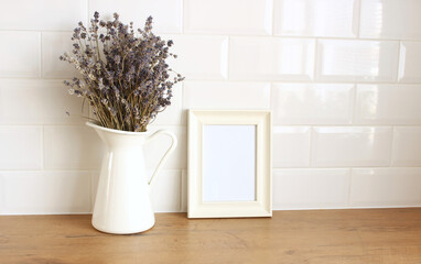 Closeup of kitchen interior. White brick wall, metro tiles, wooden table. Portrait empty wooden frame mockup with modern vase with lavender. Modern Scandinavian design.