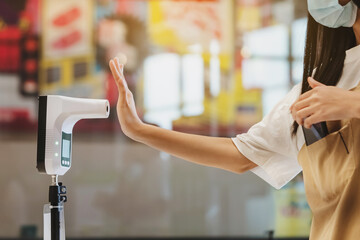 Customer with protective mask raised hand at digital thermometer with temperature scanning machine for fever before entering area. Screening to prevent infected people from entering restricted areas.