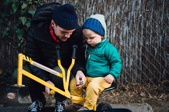 Baby boy sitting on toy excavator being repaired by crouching father