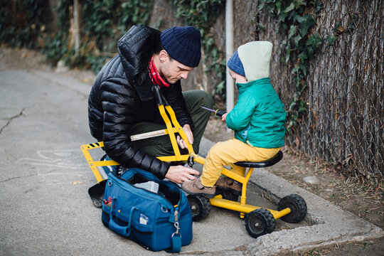 Baby boy sitting on toy excavator being repaired by crouching father