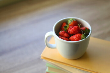 Mug filled with fresh strawberries and stack of book on a table. Selective focus.