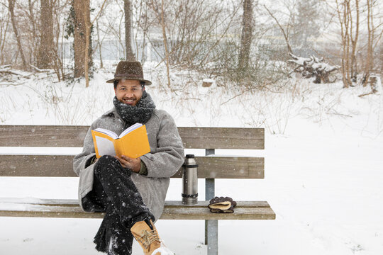 Smiling man reading book while sitting on bench during winter