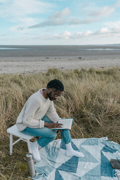 Young man reading book while sitting on stool at beach