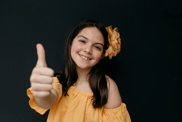 Cute girl with flower on head showing thumbs up gesture against black background