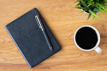 Top view of black closed cover blank notebook with coffee cup and pen for taking write notes on wood table background. Flat lay, creative workspace office. Business-education concept with copy space.