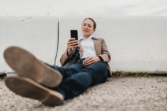Relaxed businesswoman surfing net while sitting at crash barrier on roadside