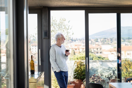 Senior man holding wineglass while leaning by sliding door at home