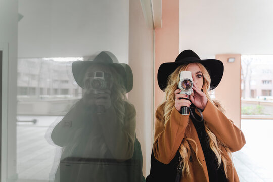 Young woman in hat filming through vintage video camera against reflection in window