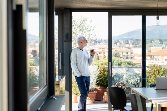 Man with wine glass looking away while standing by sliding door at  home