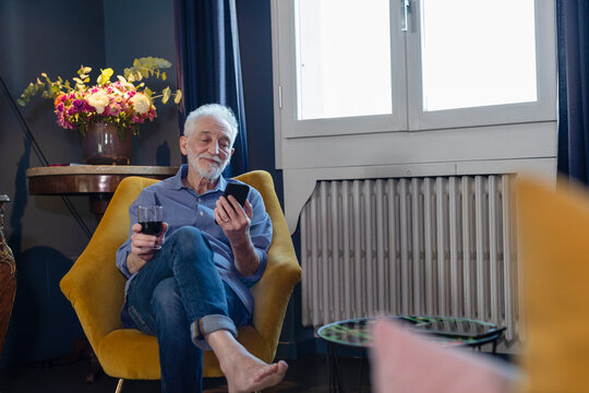 Senior man with wineglass using mobile phone while sitting on chair at home