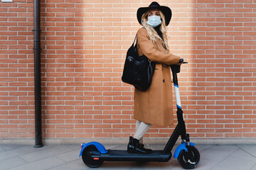 Blond woman in protective face mask with electric push scooter standing against brick wall