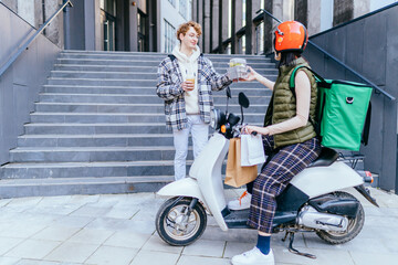 Female courier with red helmet delivering fresh lunches to young business man on a scooter with green thermal backpack. Takeaway restaurant food delivery service concept.