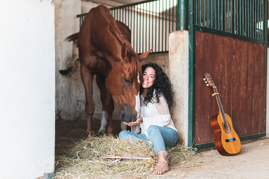 Smiling woman with guitar feeding horse while sitting in stable