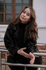 Smiling young woman in black leather jacket standing in front of white wall, leaning on iron railings, looking at camera. Urban style and street fashion. Girl with curly hairstyle in casual outfit