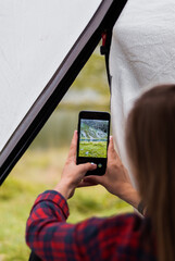 Girl taking photos of the rocky mountains and lake using the phone. Close-up shot of the handі holding the phone. View from the tent.