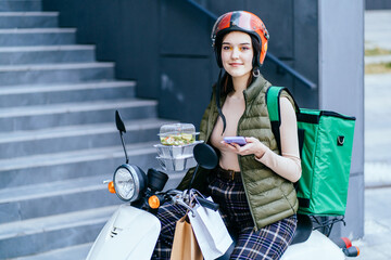 Young female courier in red helmet delivering food on a scooter, checking order with a smart phone while standing behind stairs on street in the city. Delivery service concept.