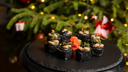 Sushi with black rice, crab meat, avocado, smoked salmon mousse, oar caviar, masago, shrimp cocktail and edible gold leaf with ginger on black table for Christmas with a Christmas tree on background.