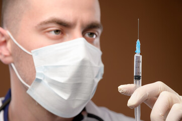 A young male Caucasian doctor in a mask holds a syringe with an injection with his gloved hand....