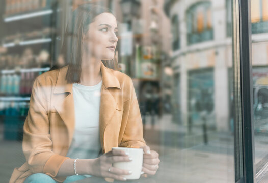 Contemplating young woman with coffee cup sitting at cafe looking through window
