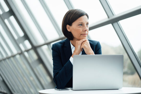 Thoughtful businesswoman with hand on chin by laptop at desk in office - Powered by Adobe