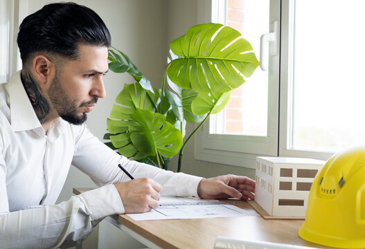 Male architect looking at architectural model while drawing blueprint in office