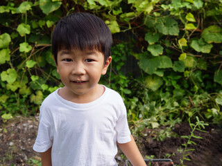 Asian cute child boy smiling with surprised face while do gardening outdoor in nature background with green leaves plant. Young kid having happy moment in summer. Concept of farming at home.