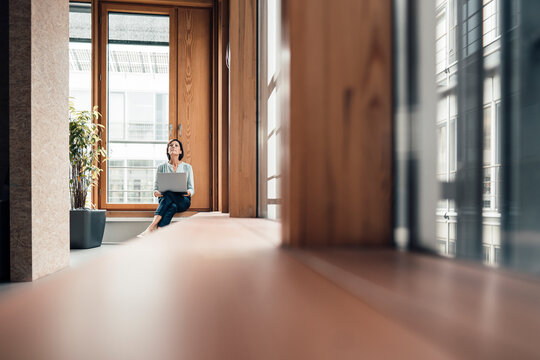 Female entrepreneur with laptop sitting on alcove widow seat at office