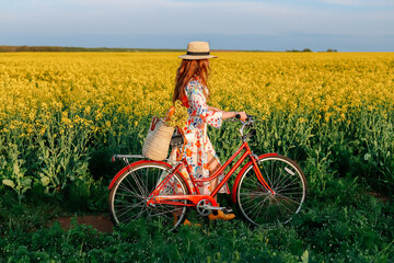 Girl with a red retro bicycle going along the yellow field of flowers.