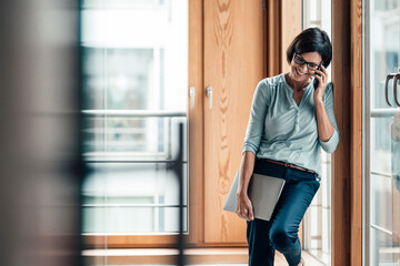 Female entrepreneur talking on smart phone while leaning over window at office