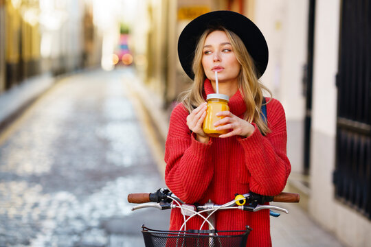 Blond woman wearing hat drinking juice while standing with bicycle on footpath