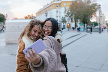 Cheerful female friends taking selfie through smart phone while embracing on sidewalk in city
