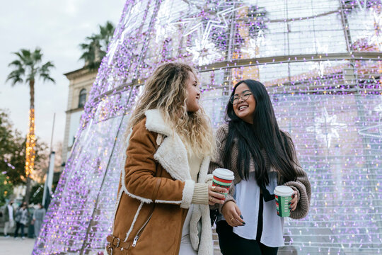 Cheerful female friends holding coffee cup while walking by Christmas decoration at street