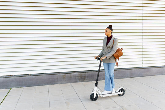 Young woman businesswoman with bag riding electric push scooter against white striped wall