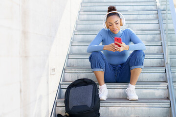 Young woman with backpack and headphones using mobile phone while sitting on steps