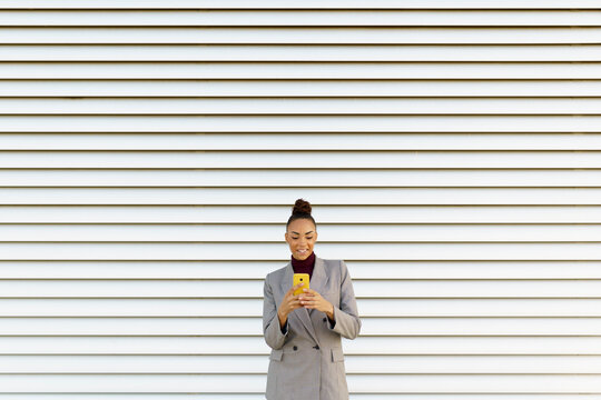 Smiling businesswoman using mobile phone while standing against white wall
