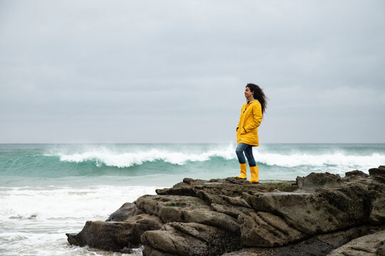 Young woman in yellow raincoat looking at sea view while standing on rock