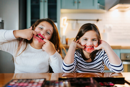 Playful girls with makeup on face teasing while sitting at dining table in domestic kitchen