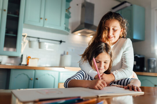 Girl coloring on paper while sister hugging from behind in kitchen