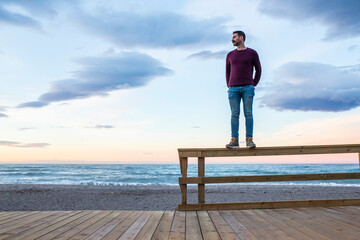 Man with hands in pockets standing on wooden railing at beach against sky